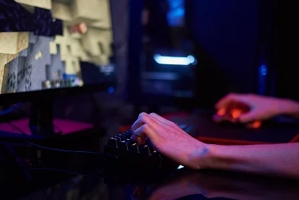 Osoba grająca na komputerze z podświetlaną klawiaturą i myszą, skupiona na ekranie, na którym wyświetla się kanciasty, pikselowy krajobraz. Przytłumiona, rozświetlona atmosfera.